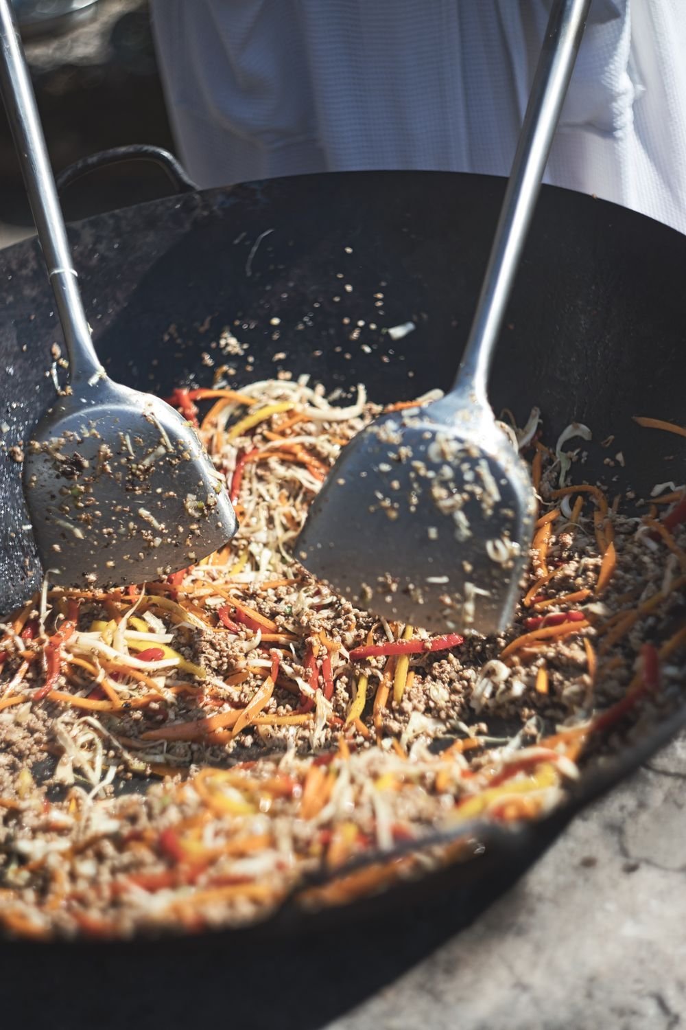 Two metal ladles tossing noodles and vegetables in a black wok