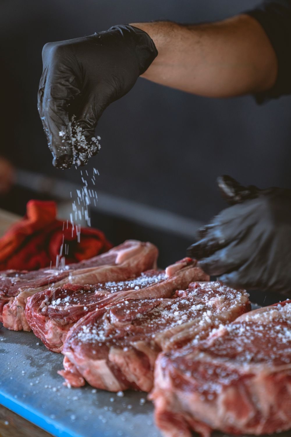 Gloved hand sprinkling coarse salt over a row of raw steaks