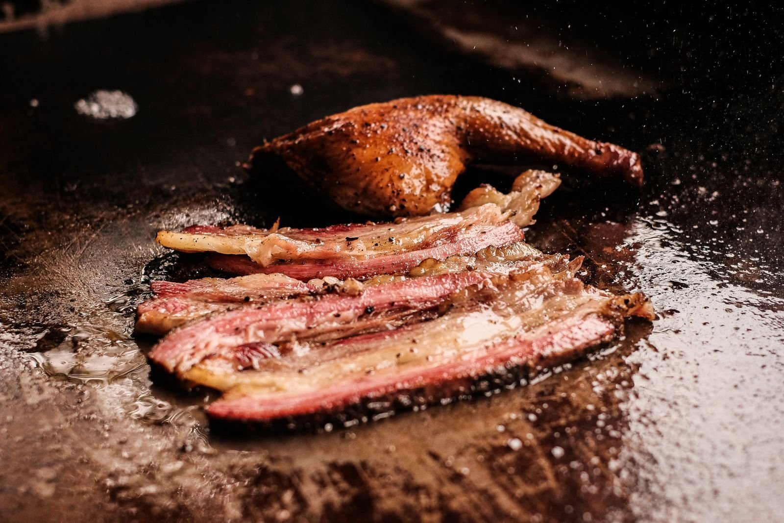 Thick bacon strips and a crisp duck leg sizzling on a griddle