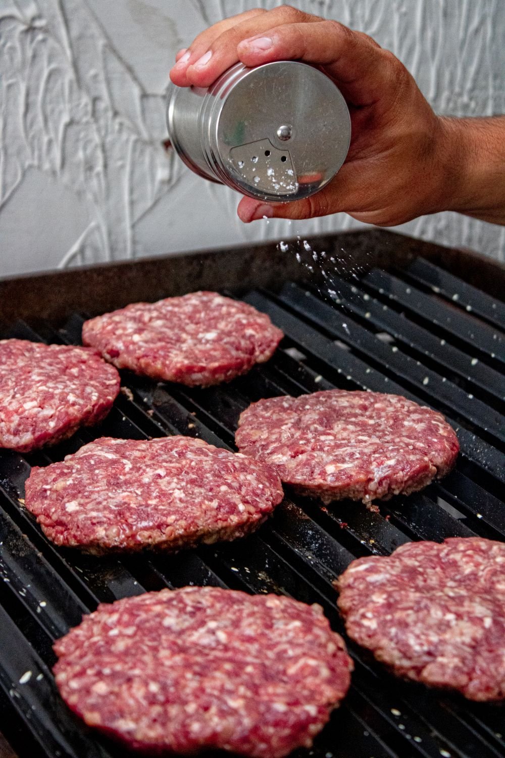 Hand seasoning raw burger patties laid out on a grill grate