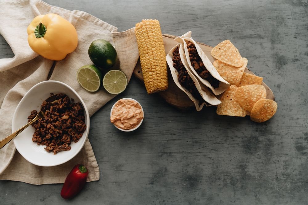 Tacos, tortilla chips, corn, limes and seasoned meat on a slate table
