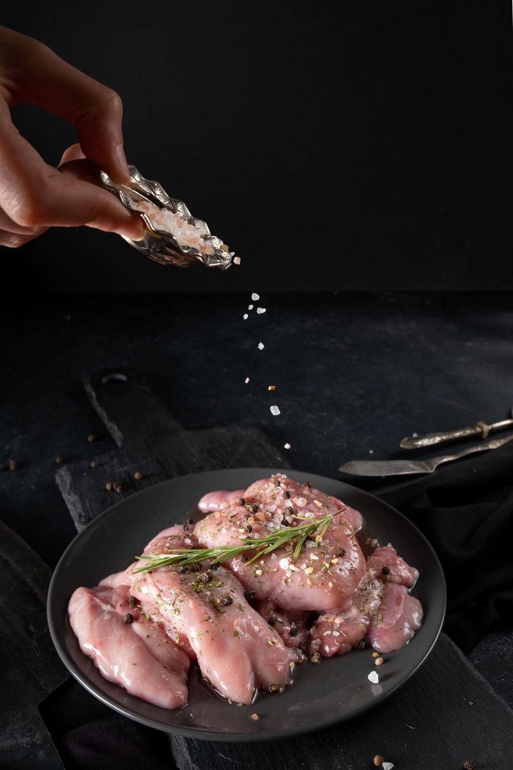 Hand sprinkling salt over raw chicken tenders in a black pan