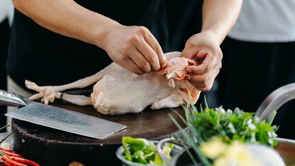 Hands trimming a whole raw chicken beside a cleaver and fresh greens