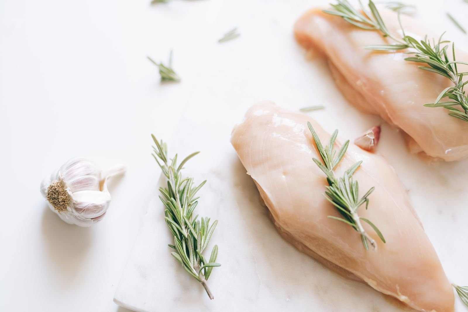 Raw chicken breasts with rosemary sprigs and garlic on white marble