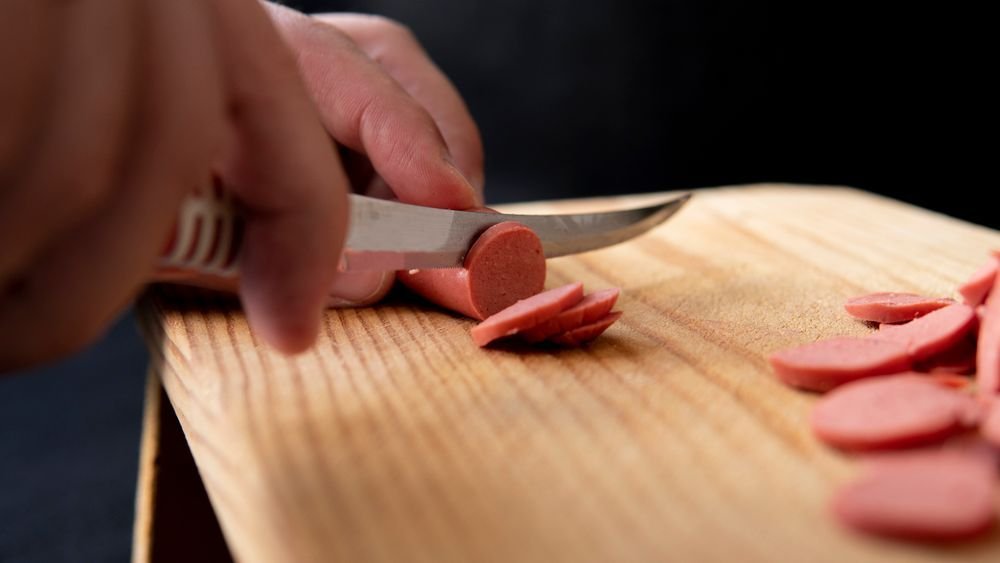 Hand cutting a sausage into thin coins on a wooden board