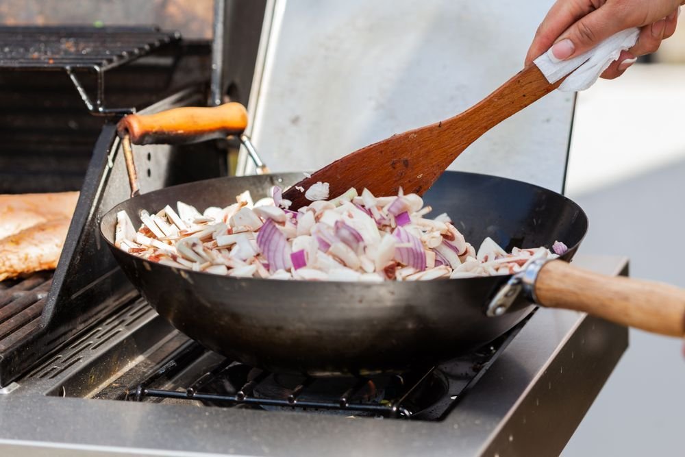 Wooden spatula stirring chopped onions in a wok set on a grill