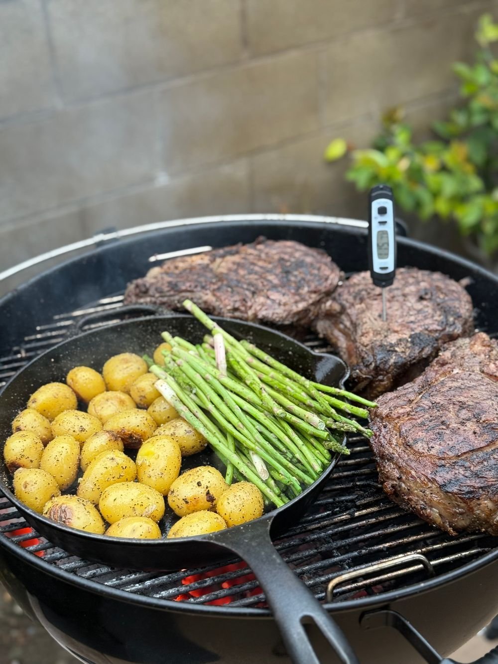 Skillet of baby potatoes and asparagus on a kettle grill beside steaks